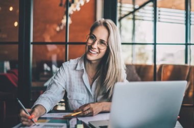 Gözlüklü, gülümseyen sarışın, kafede çalışırken masada dizüstü bilgisayar ve kağıt çizelgelerle oturuyor ve başka tarafa bakıyor.