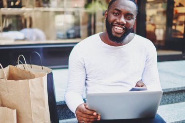 Modern akıllı telefon ve laptop cihazlarıyla merdivenlerde oturan mutlu zenci hipster portresi, neşeli Afro-Amerikan alışveriş koliği alışverişin ardından heyecanlanıyor, teknoloji kullanıcısı.