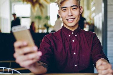 4G interneti kullanarak akıllı telefon cihazının profilini güncellerken gülümseyen Çinli pozitif hipsterin portresi. Masaüstünde oturan yakışıklı Asyalı öğrenci cep telefonu tutuyor.