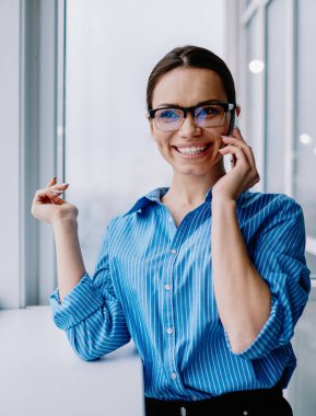 Başarılı bayan girişimci, akıllı telefondan iyi haberler almaktan mutlu. Resmi giysiler içinde ofiste duruyor. Gözlüklü neşeli bayan çalışan kariyer refahını kutluyor ve konuşuyor.