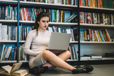 Dizüstü bilgisayar ve açık kitaplarla çalışan kendine güvenen bir kadın kütüphanede bacak bacak bacak üstüne atmış bir şekilde oturuyor.