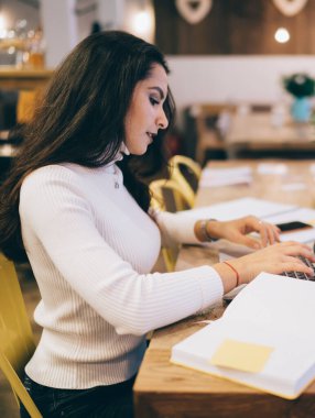 Odaklanmış, uzun siyah saçlı, beyaz kazaklı, laptoplu, renkli çıkartmalı, modern iş yerinde not defteriyle masada oturan zeki bir kadın.