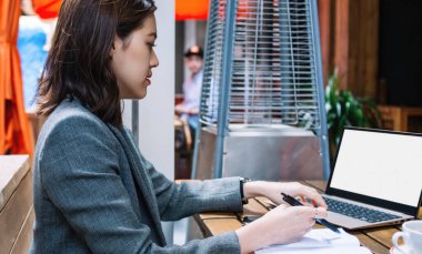 Asyalı genç bir kadının boş ekranlı dizüstü bilgisayar kullanırken ve kafede otururken not alırken yan görünümü