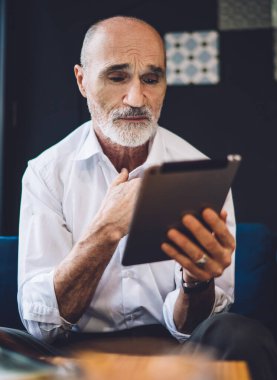 Stresli, kel bir adam resmi kıyafetler giymiş, kafeteryada elleri göğsünde oturan, hüzünlü bir ifadeyle modern tablet kullanıyor.