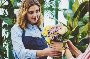 Profesyonel kadın bahçıvan kendi botanik serasından taze çiçek satıyor, önlüklü satıcı müşteriye iş sırasında cam evde bitkiler yetiştirirken ipek buket sipariş ediyor.