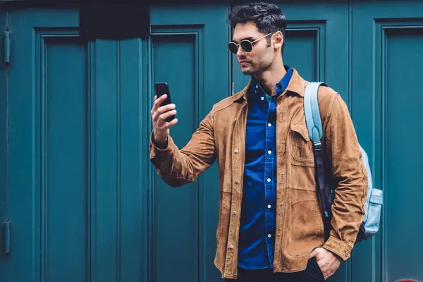 Young stylish calm male in casual clothes and sunglasses taking photo using modern digital smartphone while standing near bright blue wall at street in city