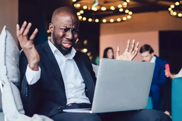 Shocked African American male office employee in formal suit raising hands with sad grimace while making mistake with application on laptop at lobby of office center
