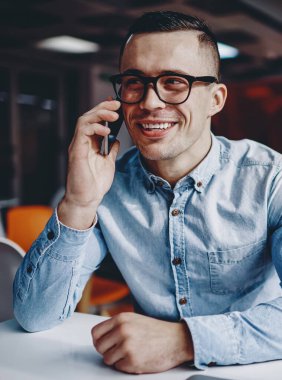 Modern çalışma ortamında masada oturan gözlüklü, akıllı telefonlu bir arkadaşla iletişim kuran pozitif hipster. Kot gömlek giymiş neşeli genç adam cep telefonundan ararken gülüyor.