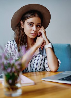 Moda şapkalı dalgın yeniyetme kız bilgisayarlı bir masada otururken kameraya bakıyor. Güzel tefekkür eden beyaz bir kadının blog yayını hakkındaki portresi.