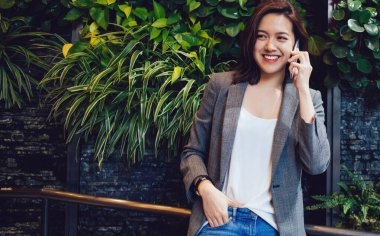 Mutlu, genç, gülümseyen, şık kıyafetler içinde, gri ceketli, mavi kotlu, telefonda konuşan güzel Asyalı kadın.