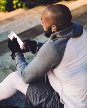 Atletik Afrikalı Amerikalı kulaklıklı ve spor kulaklıklı adam akşamları taş merdivenlerde otururken sörf cep telefonu takıyor.