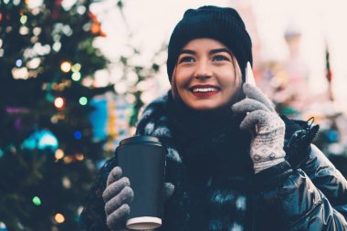 Rahat kışlık giysiler içinde mutlu bir genç kadın akıllı telefon kullanıyor ve bulanık arka planda, Noel sokağında sıcak kahve içiyor.