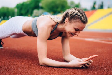 Sarışın, at kuyruklu çekici ve neşeli bir bayan stadyumda tahta örgü örüyor ve sosyal medyada bulanık arka planda gezinirken gülümsüyor.