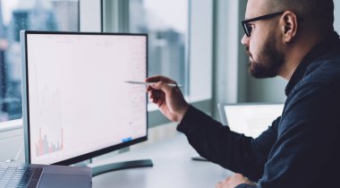 Ofisteki masadaki istatistiksel bilgilerle çalışan, gözlüklü sakallı adamın ekran görüntüsü.