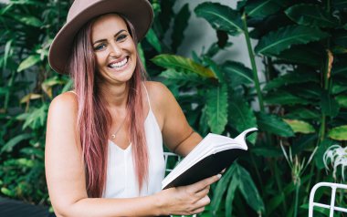 Çekici, şapkalı, saçları boyalı, kitap okuyan, tropik çalıların yanındaki masada oturan ve kameraya bakan neşeli bir kadın.
