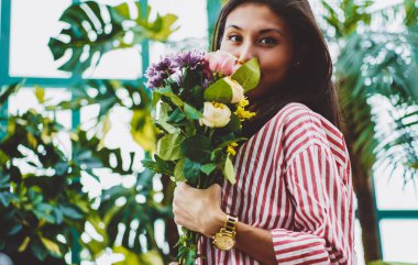 Çekici Asyalı çiçekçi kameraya bakıyor ve elinde bir buket narin aster ve gül tutuyor, cam evde elinde çiçeklerle poz veren genç satıcı, çiçek açan kompozisyon konsepti...
