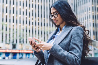Deri çanta ve cep telefonuyla şehrin arka planında duran mesajlar taşıyan gözlüklü ve modaya uygun ceketli modern etnik bayan.