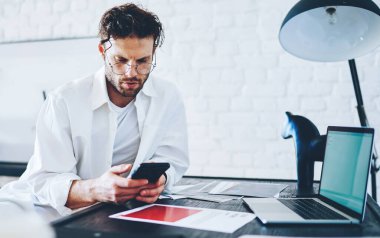 Ciddi Kafkas erkek serbest bankacı evden çalışan cep telefonundaki bakiyeyi kontrol ediyor, apartman dairesinde hippi adam uzaktan işlem yapmak için akıllı telefon kullanıyor.