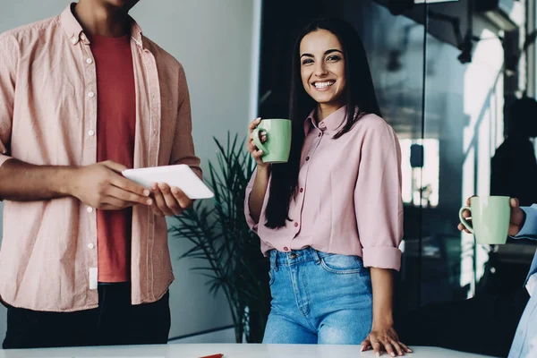 Portrait of cheerful female employee smiling a camera during coffee break with male colleagues, happy men and woman laughing during work day in office standing near desktop and using touch pad