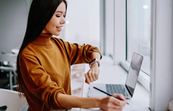 Positive brunette female office worker checking time on wristwatch while making notes near window, prosperous businesswoman satisfied with modern electronic device on wearable computer on job