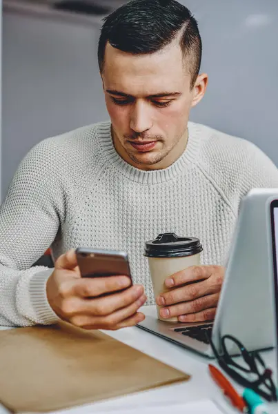 Pensive young man holding tasty coffee to go in hand and chatting online in social networks on modern smartphone using free 4G internet sitting at desktop with laptop computer with blank screen
