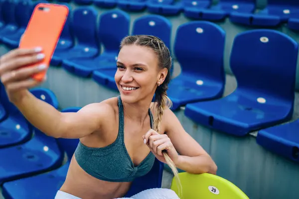 Yukardan gülümseyen, modern sportif formda yetişkin bir kadın, stadyumdaki tribün sandalyesinde otururken akıllı telefonlardan selfie fotograflarını çekiyor.