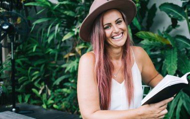 Çekici, şapkalı, saçları boyalı, kitap okuyan, tropik çalıların yanındaki masada oturan ve kameraya bakan neşeli bir kadın.