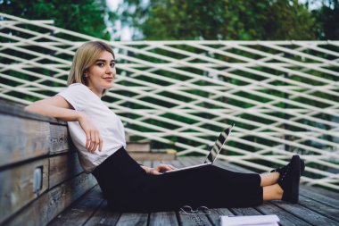 Sıradan kıyafetli genç bir kadının kitap ve dizüstü bilgisayarla ahşap tribünde oturup kameraya bakarken yan görüntüsü.