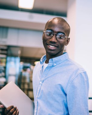 Kütüphanede duran neşeli Afro-Amerikalı adamın yan görüntüsü. Dosyası bulanık arka planda, sakallı, genç, kızıl saçlı meslektaşına gülümsüyor.