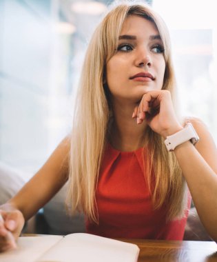 Genç sarışın kadın rahat bir kafede oturmuş, açık bir planlama içinde notlar alırken başka tarafa bakıyor ve düşünüyor.