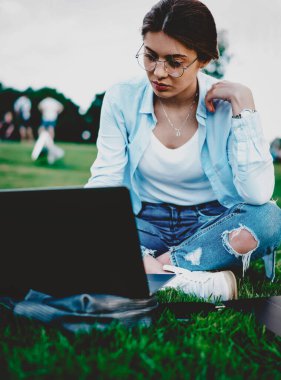 Gözlüklü genç bir kadın internet sitesinde ders seyrediyor ulusal parktaki dizüstü bilgisayarla yeşil çimlerde oturuyor. Modern netbook 'ta bilgi arayan bir öğrenci.