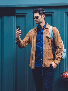 Gündelik giysiler ve güneş gözlüğü takmış, modern dijital akıllı telefon kullanarak fotoğraf çeken genç, sakin bir erkek şehrin göbeğinde parlak mavi bir duvarın yanında dikiliyor.