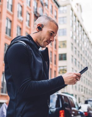 Siyah spor ceket giyen ve kulaklık takan etnik bir adamın şehrin caddesinde telefon kullanarak uygulamadan müzik seçerken yan görüntüsü