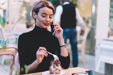 Pembe saçlı, şık giyinmiş, şık bir kadın akıllı telefondan sohbet ediyor ve hipster kafeteryasında oturmuş tatlı yiyor.