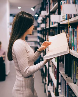 Kitap rafının önünde duran ve elinde kitap tutan güzel, ince bir kadının yan görüntüsü.