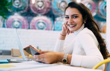 Neşeli bir kadının cep telefonuna göz atması, dizüstü bilgisayar ve not defteriyle ders çalışması gündüz şık bir kafeteryada oturup kameraya bakması.