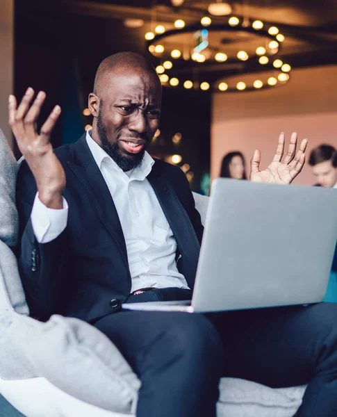 Shocked African American male office employee in formal suit raising hands with sad grimace while making mistake with application on laptop at lobby of office center