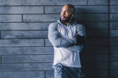 Beyaz kulaklıklı, beyaz kulaklıklı, duvara yaslanmış ve kollarını kavuşturarak fotokopi çekilen ve müziğin keyfini çıkaran kendine güvenen kel, kel sporcu.