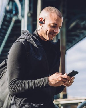 Sırt çantalı, siyah spor elbiseli, telefon kullanan ve köprü altında müzik dinleyen neşeli genç bir erkek.
