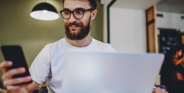 Gözlüklü, gülümseyen hippi dizüstü bilgisayarda çalışırken akıllı telefondan e-postaları kontrol ediyor. Kablosuz bağlantı paylaşımından memnun olan pozitif adam, cep telefonuyla birlikte çalışan bir yerde multimedya paylaşıyor.