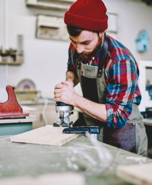 Kareli gömlekli, kırmızı sıcak şapka ve önlüklü düşünceli işçi. Konsantre oluyor ve ışık çağındaki atölyede çalışırken kalas kesmek için elektrikli yapboz kullanıyor.