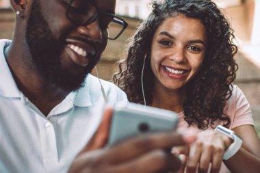 Milenyum erkek arkadaşıyla açık havada oturan cep telefonu cihazıyla halka açık sitede popüler müzik eşliğinde yeni bir podcast ararken kameraya gülümseyen esmer tenli kadının portresi