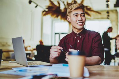 Sırıtkan Çinli hippi öğrenci dizüstü bilgisayarla masaüstünde oturuyor ve modern uzayda bayan meslektaşlarıyla birlikte iş kurma projesinde işbirliği yapma planını tartışıyor.