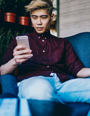 Modern akıllı telefondan 4G internete bağlı takipçilerden gelen tebligatı okuyan düşünceli Çinli hipster blogcusu modern uzay sandalyesinde oturuyor. Japon genç adam cep telefonuyla video izliyor.