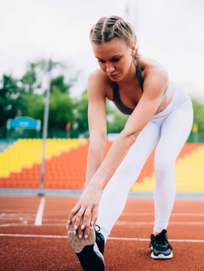Spor giyimli, sarışın, aktif genç bir kadın yazın stadyum pistinde ayak parmaklarına yaslanırken bacak kaslarını esnetiyor.