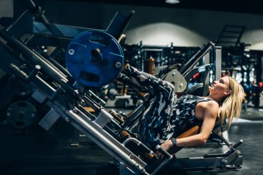 Sıkı şehir kıyafetleri ve spor ayakkabıları içinde güçlü sarışın vücut geliştirici modern spor kulübünde bacak pres makinesiyle ağırlık kaldırıyor.