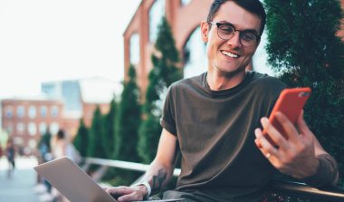 Görme düzeltmesi için gözlüklü neşeli erkek öğrenci akıllı telefon cihazıyla gülerek kampüste modern laptopla otururken, dışarıda serbest çalışan pozitif bir serbest oyuncu tarafından komik bir mesaj aldı.