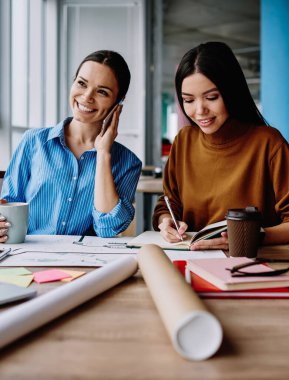 Masaüstünde birlikte tasarım yapmayı planlayan başarılı kadın ofis çalışanları. Akıllı telefondan arayan beyaz bir kadın. Neşeli meslektaşı not defterine bilgi kaydediyor.