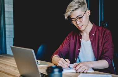 Gözlüklü zeki Asyalı genç adam dizüstü bilgisayarda webinar izlerken metin bilgileri yazıyor. Japon hippi öğrenci modern çalışma alanında dijital bilgisayarın başında ödev yapıyor.