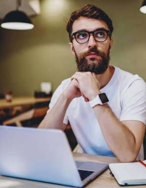 Gözlüklü, düşünceli, beyaz bir erkek birlikte çalışma alanındaki netbook 'ta uzaktan kumandayla çalışarak yayınlama fikirleri üzerinde kafa yoruyor. Düşünceli hipster adam, kafeteryadaki çevrimiçi projeye yoğunlaştı.